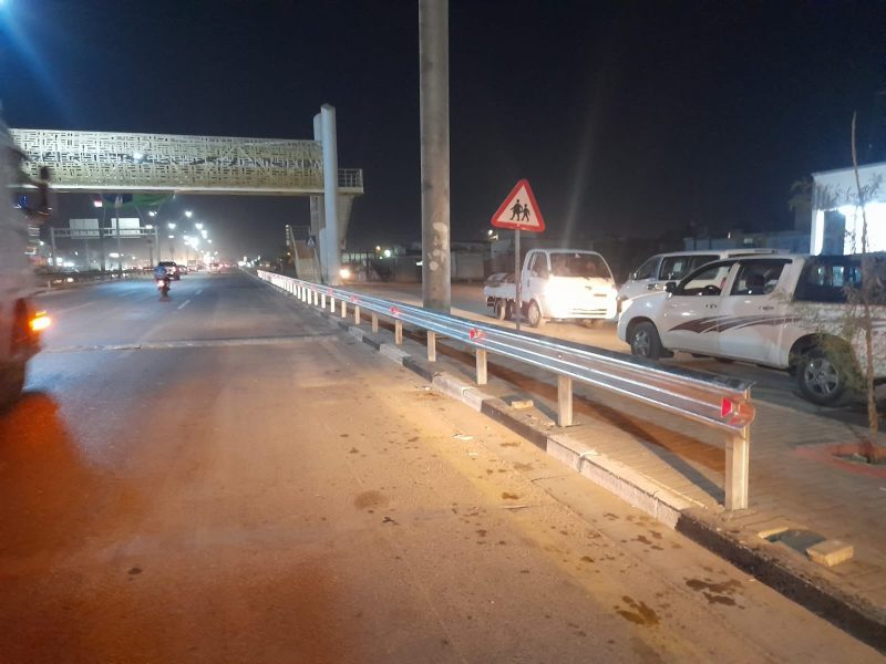 Completed W-beam median guardrail with reflective delineators; finished footbridge in the background at night.