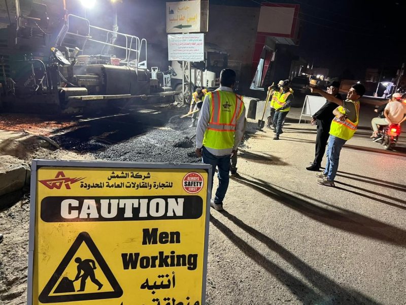 Night paving under full traffic management with bilingual "Caution — Men Working" signage.